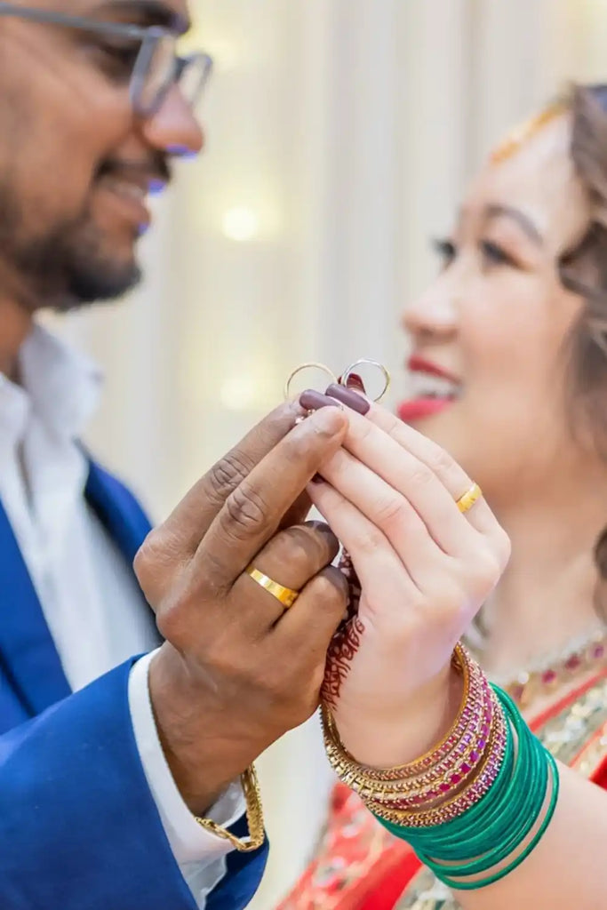 Two hands wearing wedding rings clasped together, with one hand adorned in traditional Indian bangles.